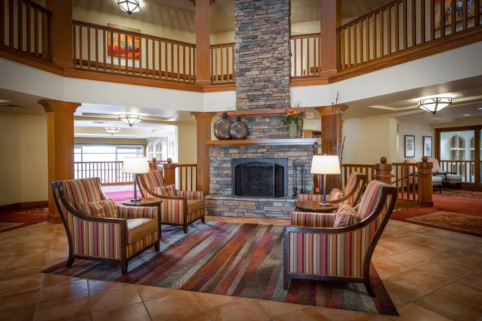 A cozy seating area in a senior living facility featuring four striped armchairs arranged around a stone fireplace. The space has warm lighting with two table lamps, a colorful area rug, wooden railings on the upper level, and decorative items on the fireplace mantel.