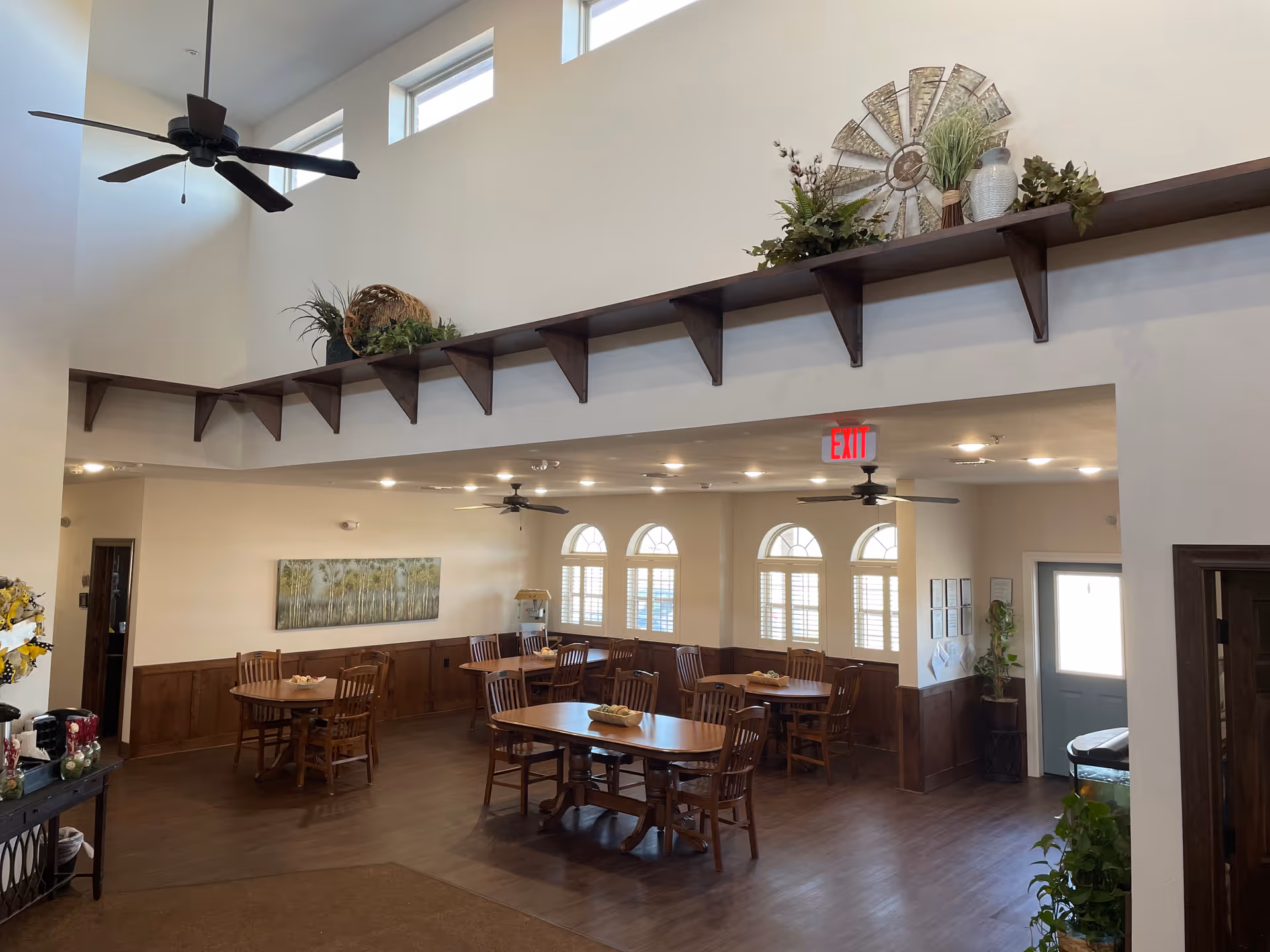 Interior view of a dining room in a senior living facility with multiple wooden tables and chairs arranged neatly. The room has high ceilings with ceiling fans, large windows with shutters, and decorative plants on a shelf above the entrance. There is an exit sign above a doorway and a painting on the wall.
