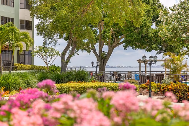 View of a landscaped outdoor area with pink flowers in the foreground, green bushes, trees, and a waterfront with a railing and outdoor seating. Part of a building is visible on the left side.
