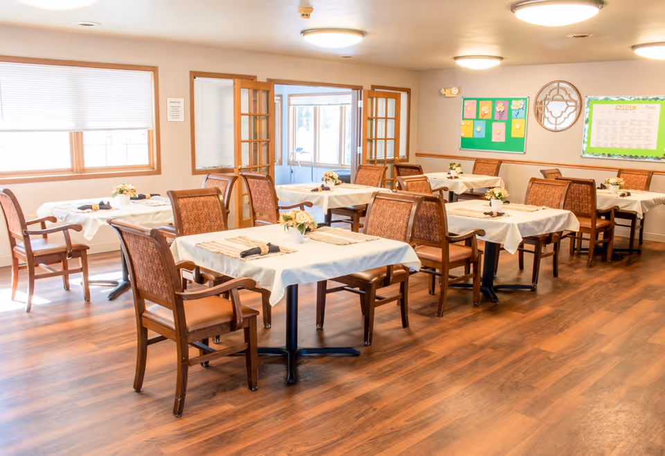 A bright dining room with several tables covered in white tablecloths, each set with placemats, napkins, and small floral centerpieces. The room has wooden floors, cushioned wooden chairs, large windows with blinds, and a bulletin board on the wall. French doors lead to another room with large windows.