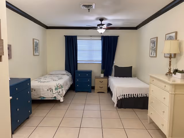 A simple bedroom with two single beds placed against opposite walls. The bed on the left has a floral bedspread, and the bed on the right has a white textured blanket with a dark bed skirt. Between the beds is a small beige nightstand with two drawers and a small plant on top. A blue chest of drawers is positioned under a window with closed blinds and dark blue curtains. On the right side of the room is a white dresser with a lamp and small plants on top. The walls are light-colored with dark crown molding, and the floor is tiled.