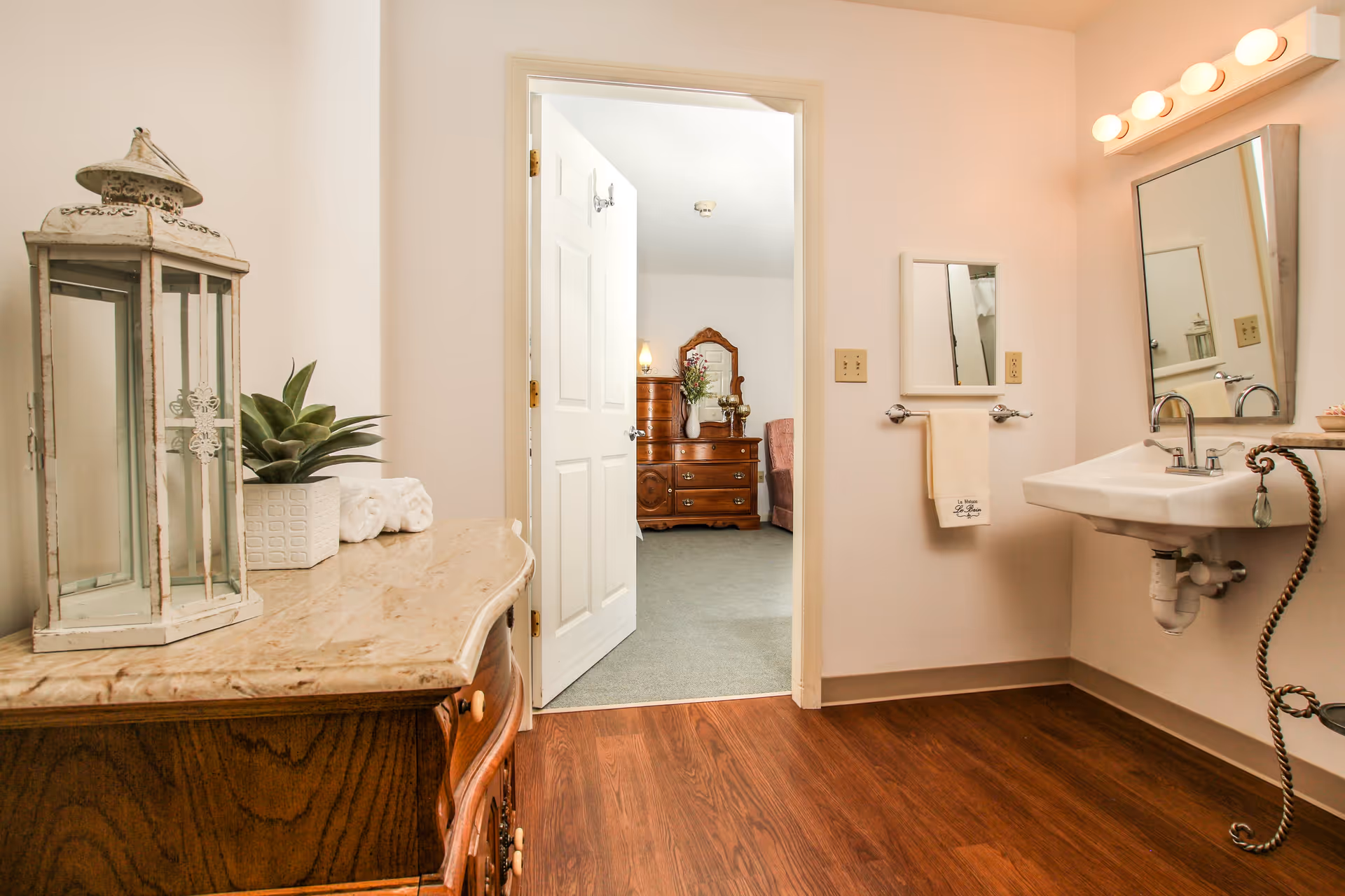 Interior view of a bathroom with a white sink, a large mirror with round lights above it, a towel rack with a beige towel, and a wooden cabinet with a marble top holding a decorative lantern and a potted plant. The bathroom door is open, showing a glimpse of a bedroom with a wooden dresser, a mirror, and a chair.