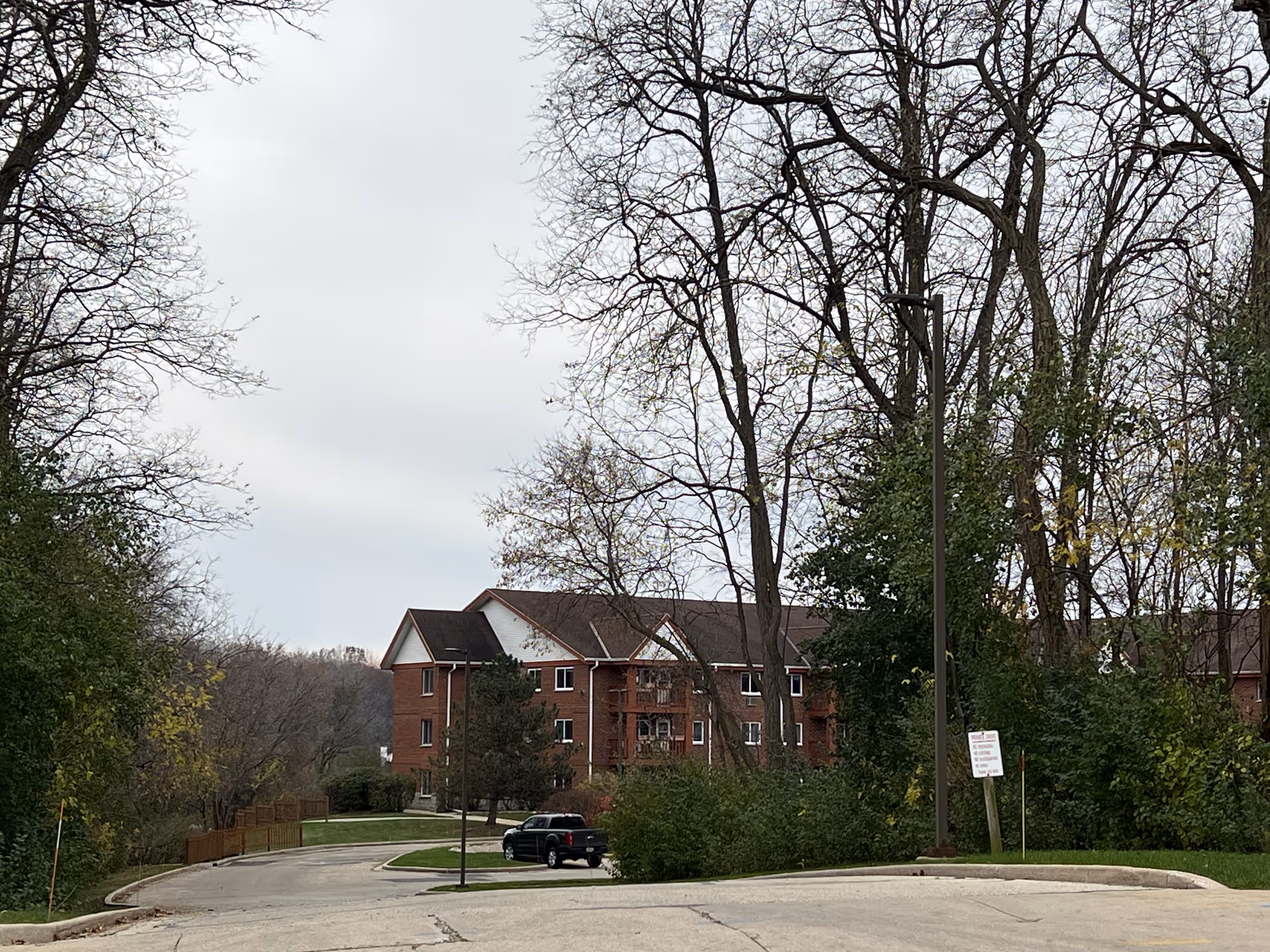 Brick multi-story senior living building seen through trees with a curved driveway and a parked car in front.