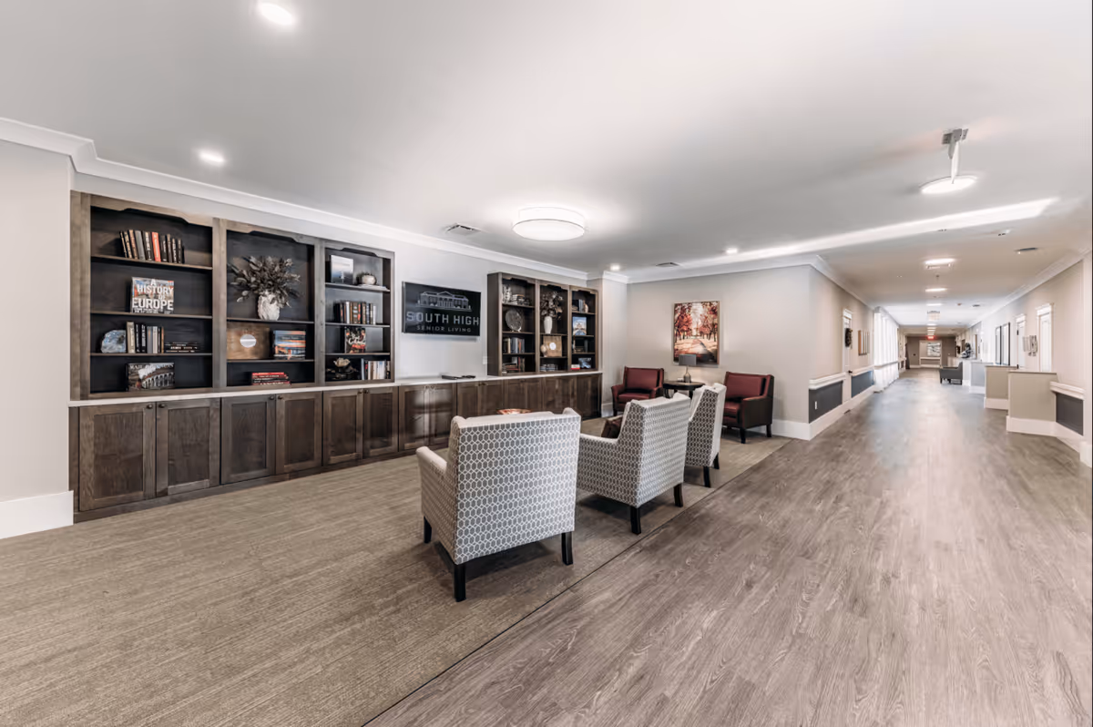 A spacious common area in South High Senior Living with built-in bookshelves, armchairs, and a long corridor.