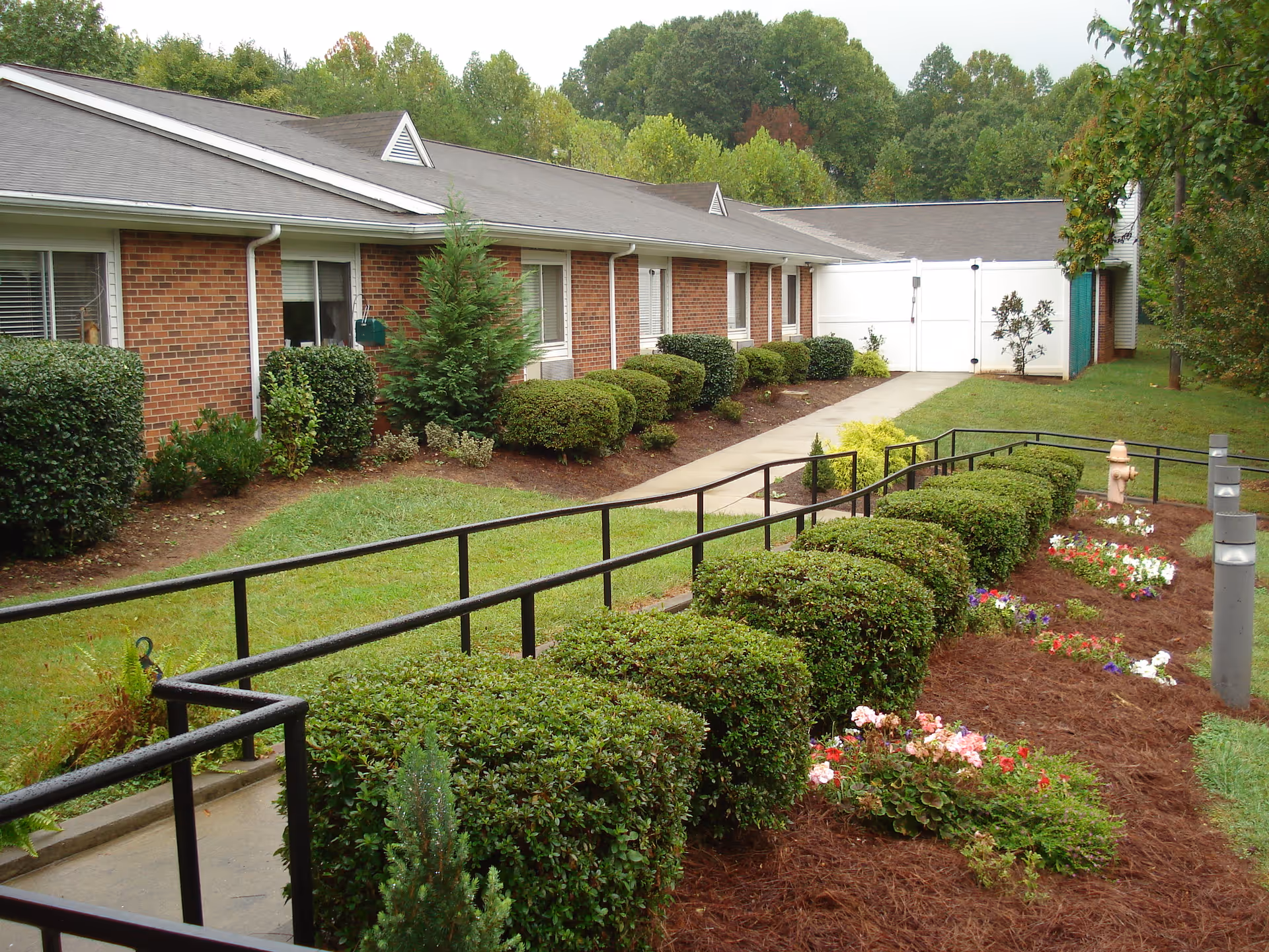 Single-story brick building exterior with a paved walkway and handrail ramp bordered by trimmed hedges and flowerbeds.
