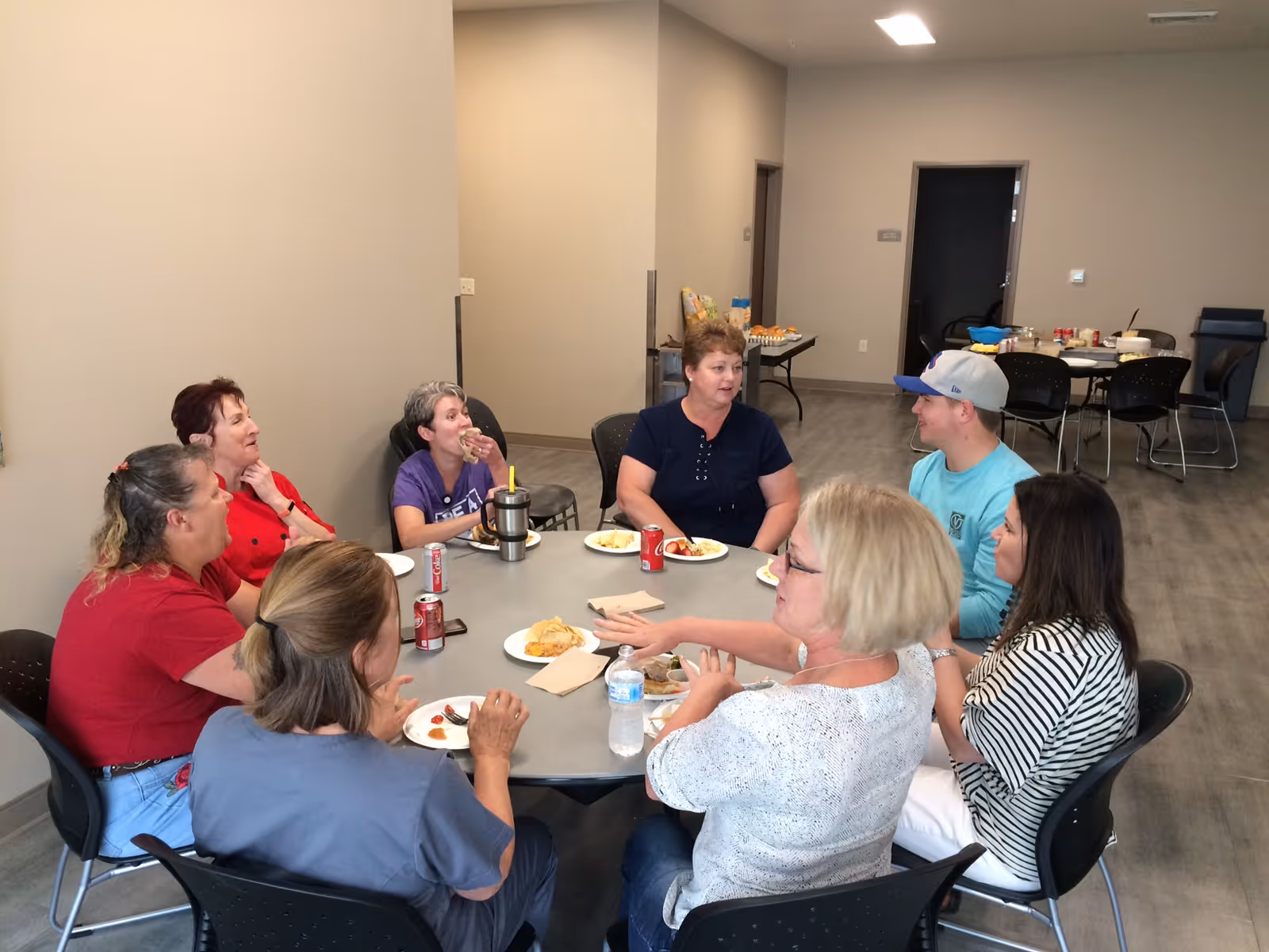 A group of seven people sitting around a round table in a room, eating and talking. The table has plates with food, soda cans, and a water bottle. The room has a plain beige wall and a gray floor, with additional tables and chairs in the background.