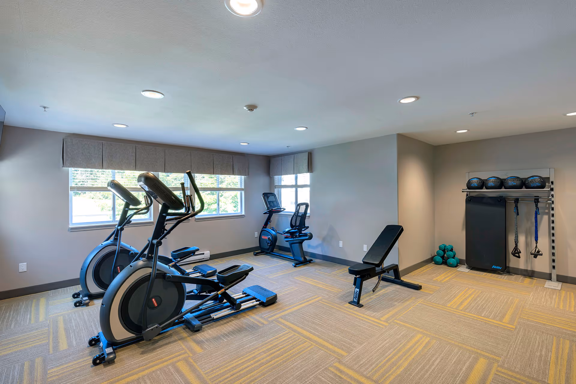 A small fitness room with two elliptical machines, a recumbent exercise bike, a weight bench, and a rack holding medicine balls, dumbbells, and resistance bands. The room has large windows with gray valances, recessed ceiling lights, and a patterned carpet with yellow and gray tones.