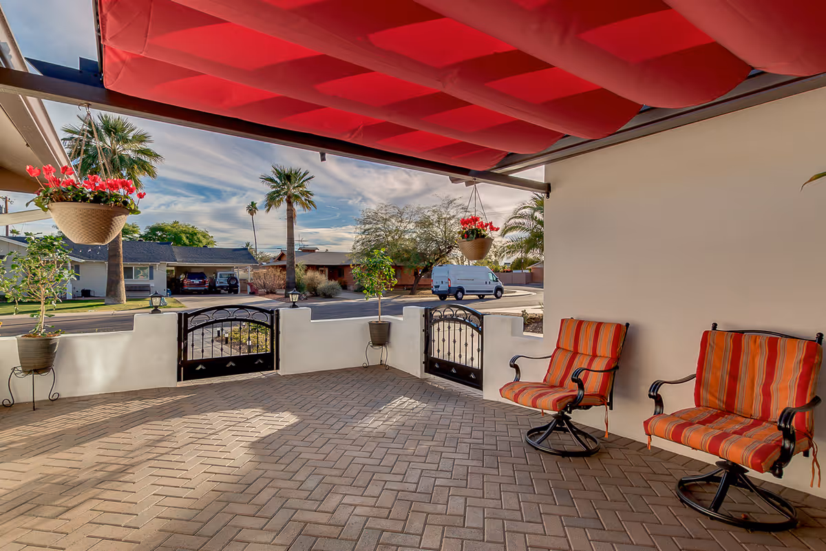 A covered outdoor patio area with two striped cushioned chairs, hanging flower pots with red flowers, potted plants, and a small black metal gate leading to a street with palm trees and houses in the background under a partly cloudy sky.