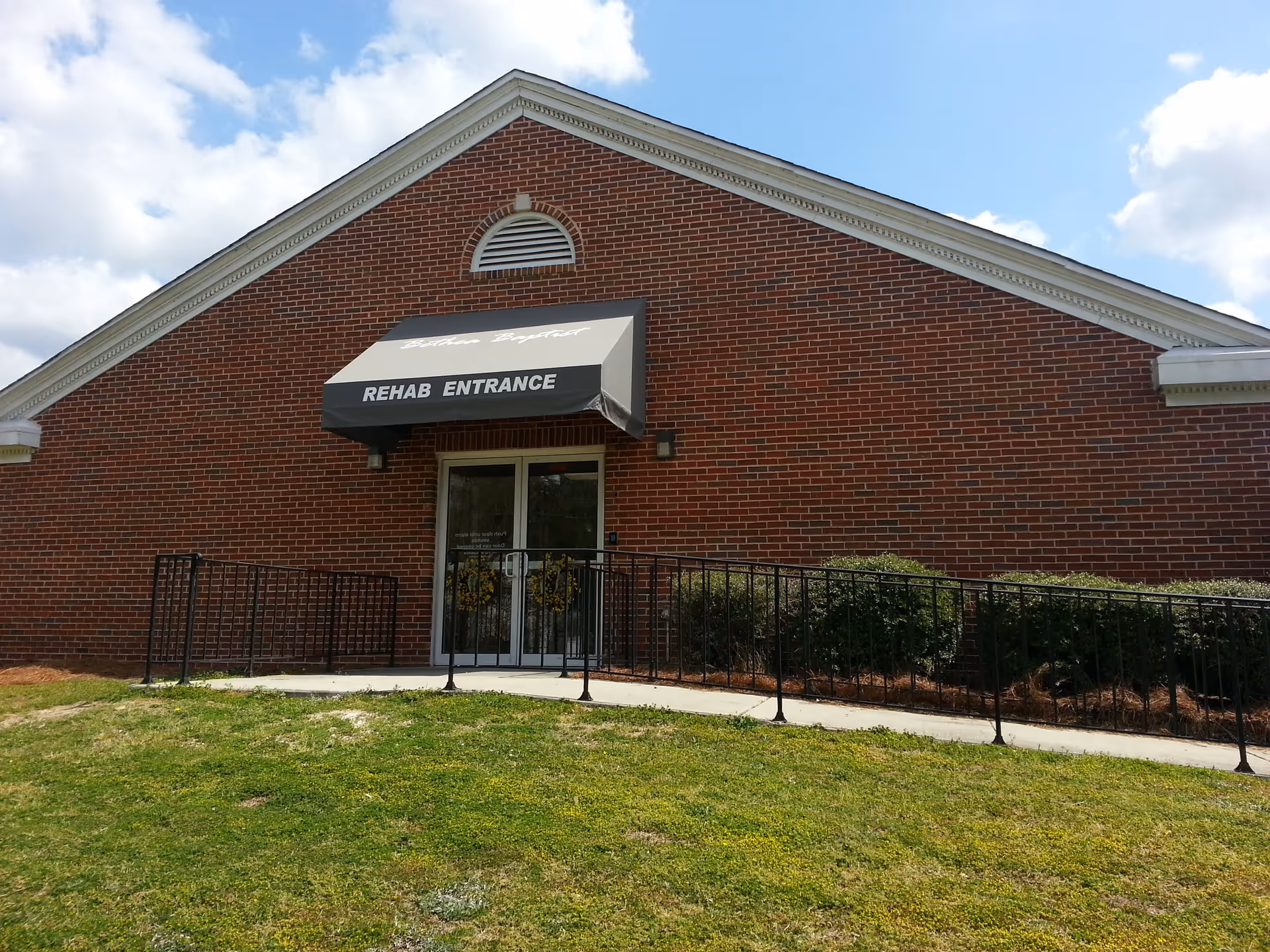 Exterior view of a brick building with a black and white awning above a glass door entrance labeled 'REHAB ENTRANCE'. There is a metal handrail ramp leading up to the entrance, surrounded by green grass and bushes. The sky is partly cloudy.