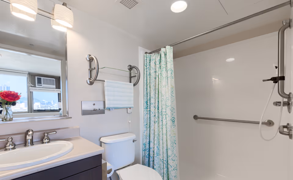 A clean and modern bathroom with a white sink and countertop, a large mirror above the sink, a toilet, and a shower area with a patterned shower curtain. The shower has grab bars for accessibility. A window with a view of buildings and a red flower in a vase is reflected in the mirror.