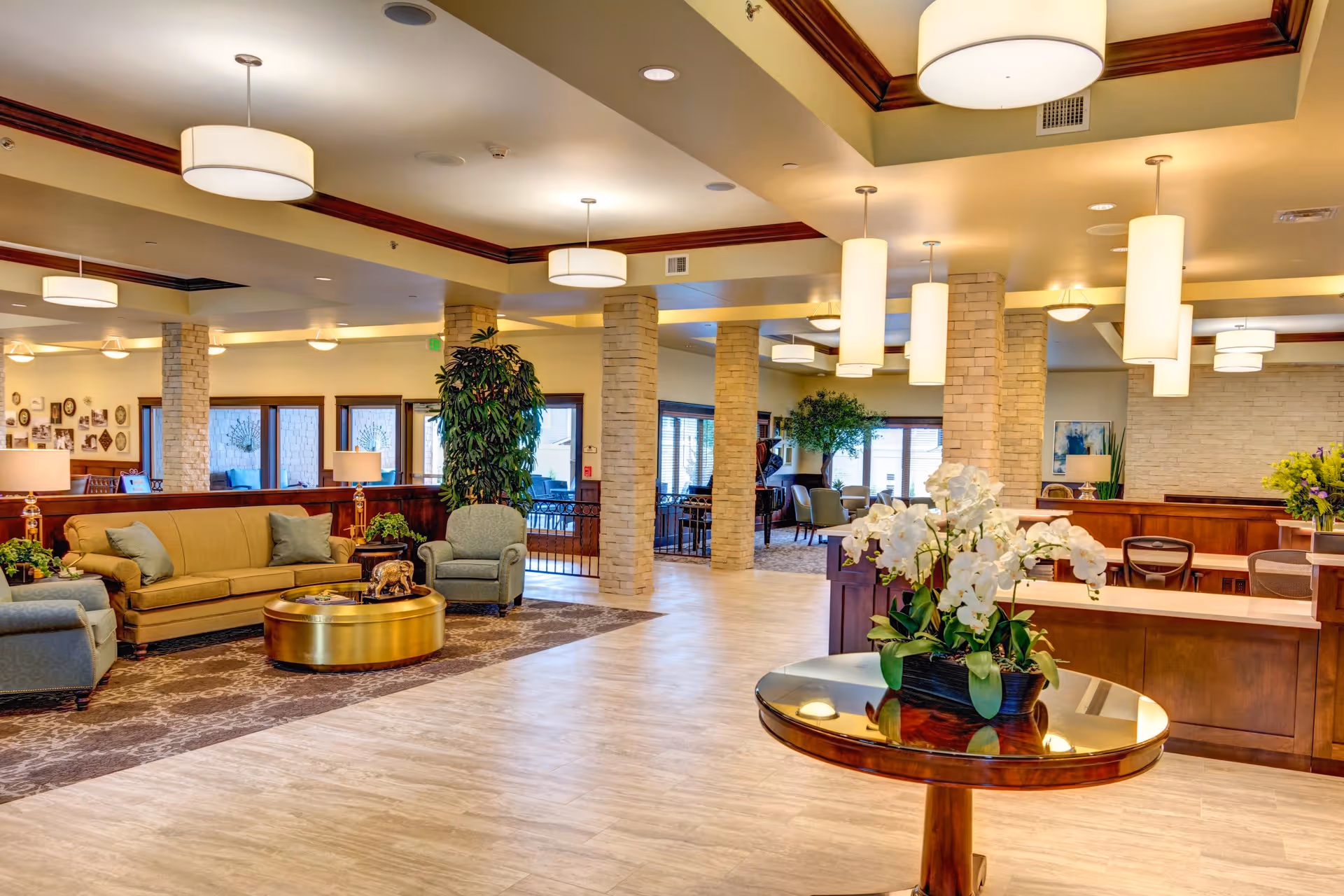 Spacious and well-lit senior living facility common area with comfortable seating including sofas and armchairs, round coffee tables, decorative plants, and modern ceiling lights. The room features light-colored flooring, stone pillars, and large windows allowing natural light to enter.