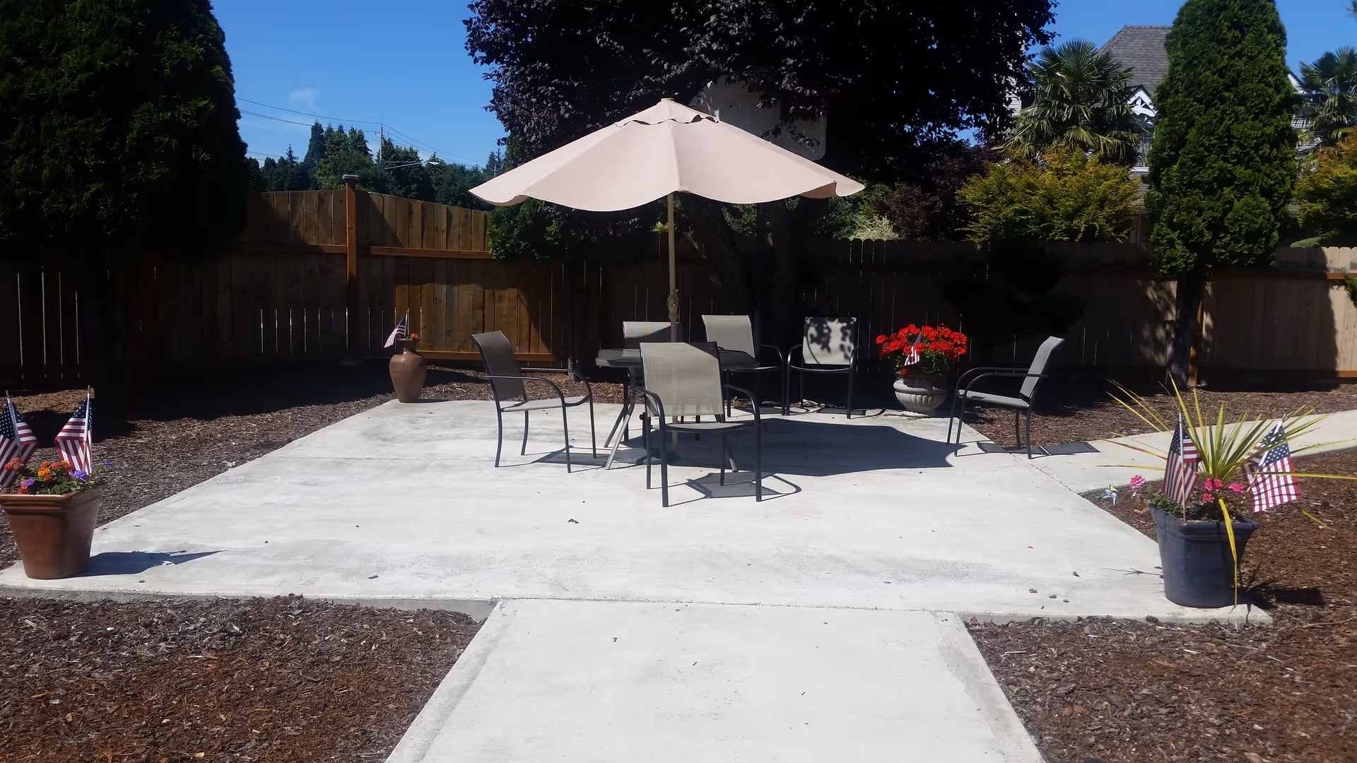 Outdoor patio area with a concrete floor, featuring a round table with a large beige umbrella and several chairs around it. The patio is surrounded by mulch beds with potted plants and small American flags. Tall trees and a wooden fence enclose the space.