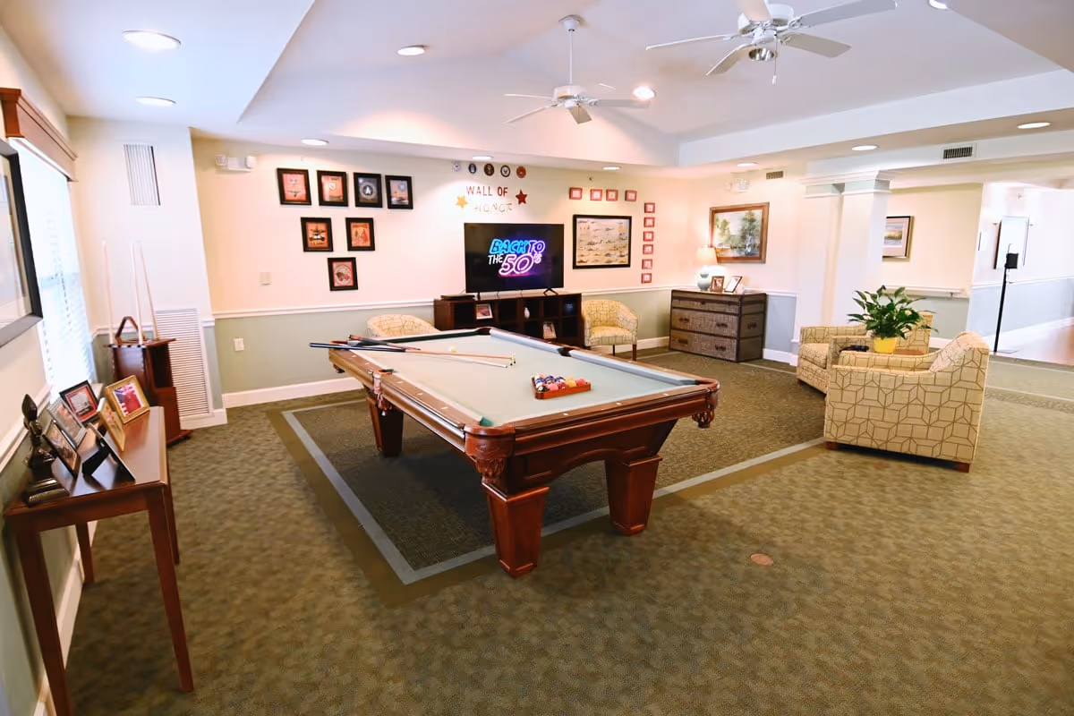 Well-lit recreation room with a pool table in the center, seating areas, a TV, and framed wall decor.
