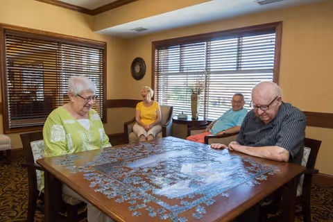 Four elderly individuals sitting around a wooden table working on a large jigsaw puzzle in a well-lit room with windows covered by wooden blinds.