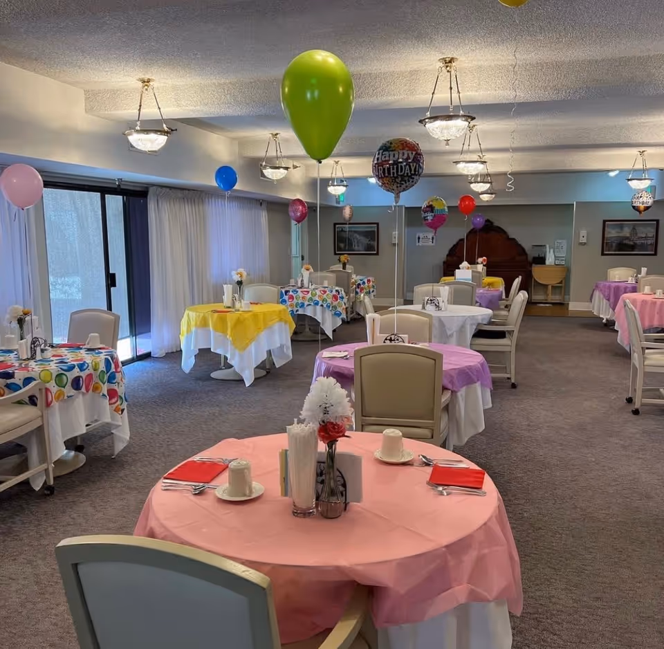 A decorated dining room with multiple round tables covered in colorful tablecloths and set with cups, plates, and utensils. Each table has a balloon tied to it, including some that say 'Happy Birthday.' The room has carpeted floors, ceiling lights, and large windows with curtains.