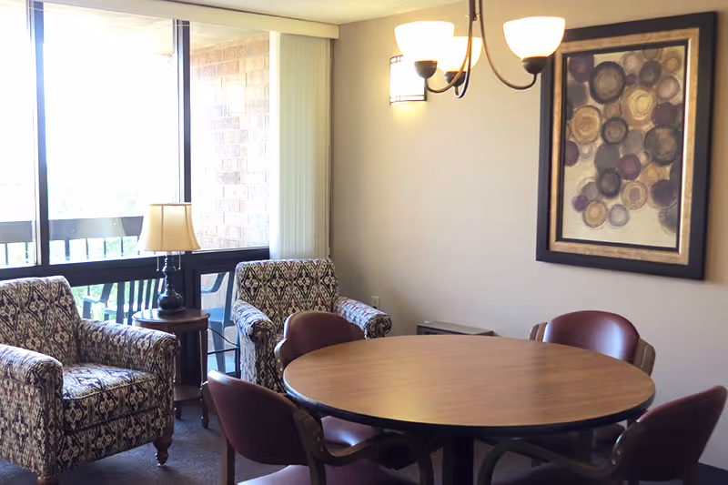 A cozy interior room with a round wooden table surrounded by four maroon chairs. Two patterned armchairs are placed near a window with vertical blinds, next to a small round side table with a lamp. A framed abstract artwork hangs on the beige wall, and a ceiling light fixture with three lamps provides illumination.