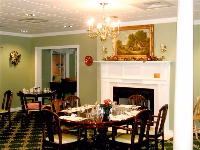 A dining room with a round wooden table set for a meal, surrounded by six chairs with cushions. The room features a white fireplace with a decorative mantel, above which hangs a framed painting of a countryside scene. A chandelier with lit bulbs hangs from the ceiling, and the walls are painted green. Additional tables and chairs are visible in an adjacent room through an open doorway.