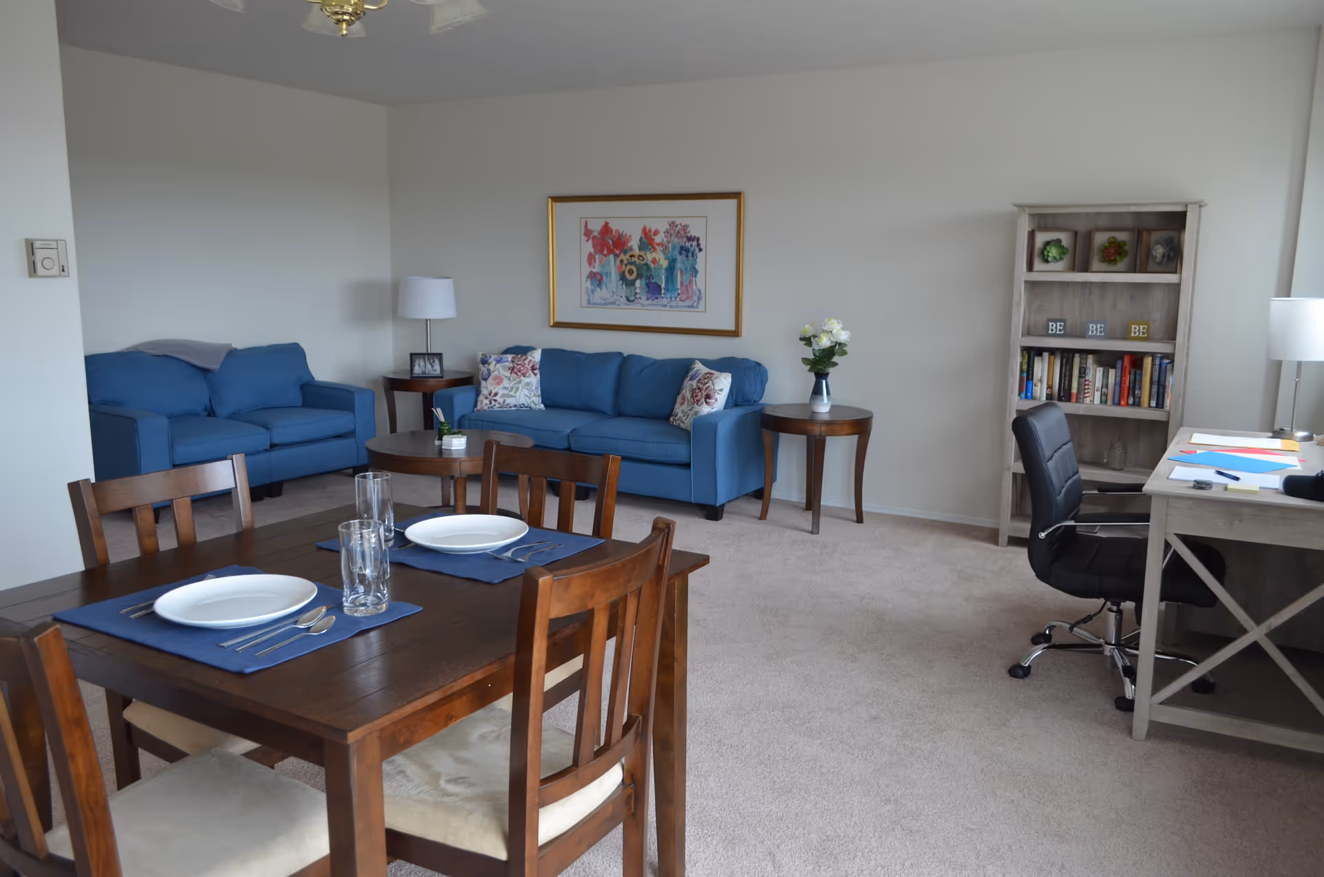 A living area in a retirement community featuring two blue sofas with floral pillows, a wooden dining table set for two with plates and glasses, a wooden coffee table, two side tables with lamps and a vase of flowers, a framed floral painting on the wall, a bookshelf with books and decorative items, and a desk with a black office chair and papers.