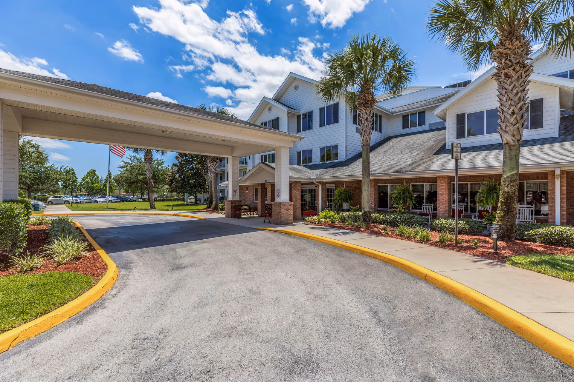 Covered entrance and driveway of a multi-story senior living building with palm trees and a porch with rocking chairs.