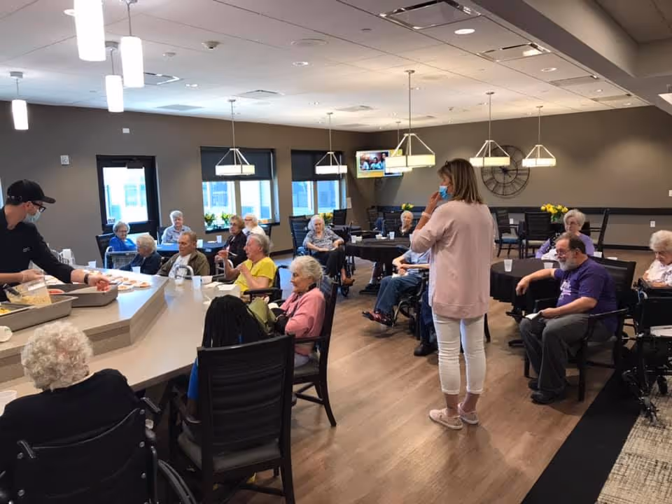A group of elderly people seated around tables and a counter in a well-lit room, with a staff member serving food and another staff member standing and wearing a mask. The room has modern lighting fixtures, large windows, and a wall clock.