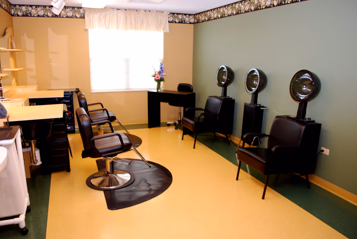 Interior salon room with styling chairs and hooded hair dryers along a green wall and workstations by a window.