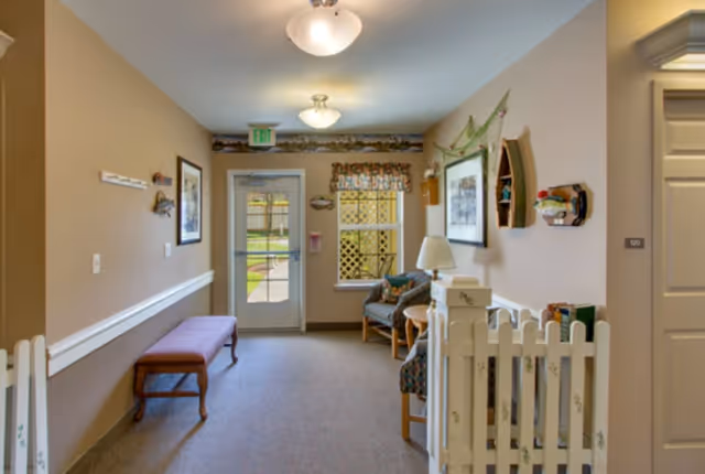 A cozy hallway in a senior living facility with beige walls and carpeted floor. There is a wooden bench on the left side, two patterned armchairs with a small round table and lamp on the right side, and a white picket fence gate near the foreground. The hallway has a door with a window and an exit sign above it, and a window with a decorative valance. Various wall decorations and framed pictures adorn the walls.