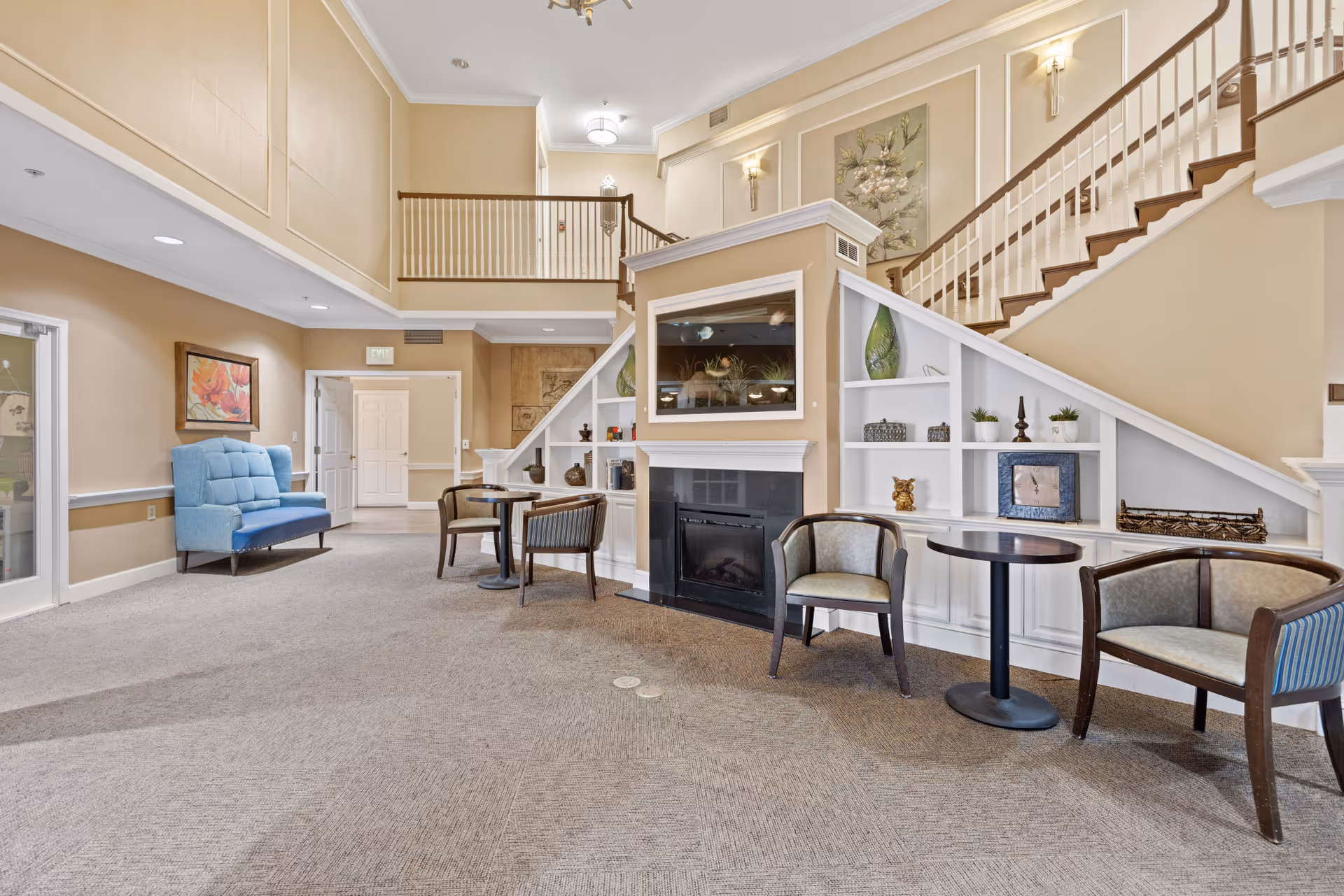 A spacious and well-lit interior common area with beige walls and carpeted floor. The room features a staircase with white railings, built-in shelves under the stairs decorated with vases, a clock, and small plants. There are several chairs and small round tables arranged near a fireplace with a mounted TV above it. A blue upholstered bench is placed against one wall, and framed artwork hangs on the walls.