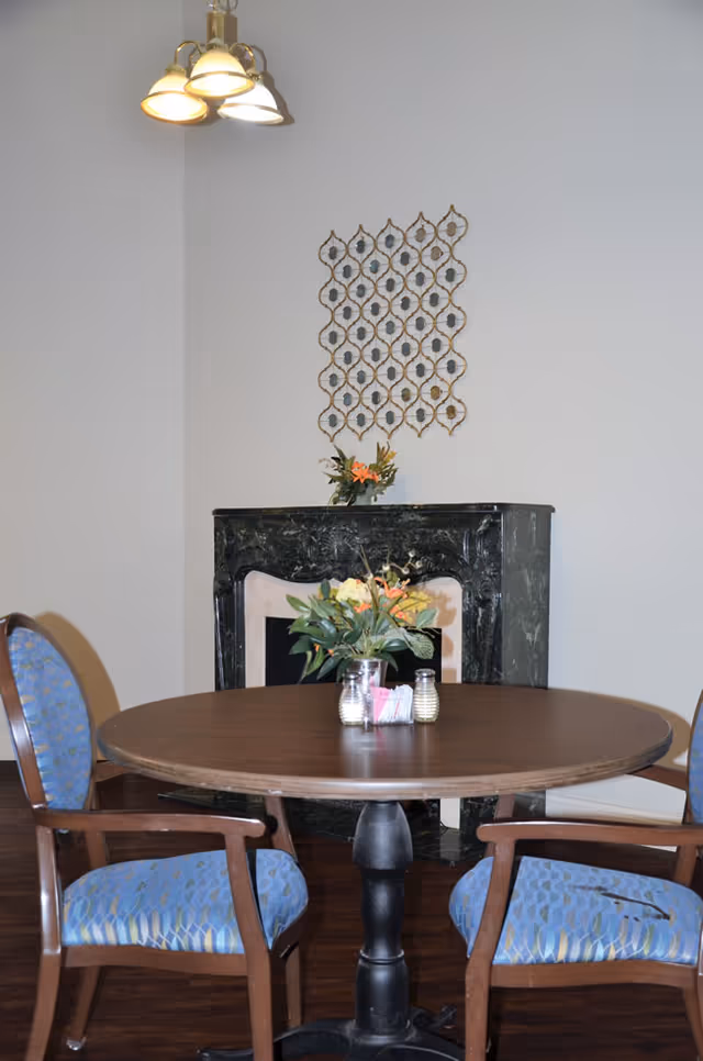 A round wooden table with a black pedestal base surrounded by two wooden chairs with blue patterned cushions. On the table, there is a small flower arrangement and salt and pepper shakers. Behind the table, there is a dark green marble fireplace with a small floral decoration on the mantel and a decorative wall hanging above it. A ceiling light fixture with four lamps is visible above.