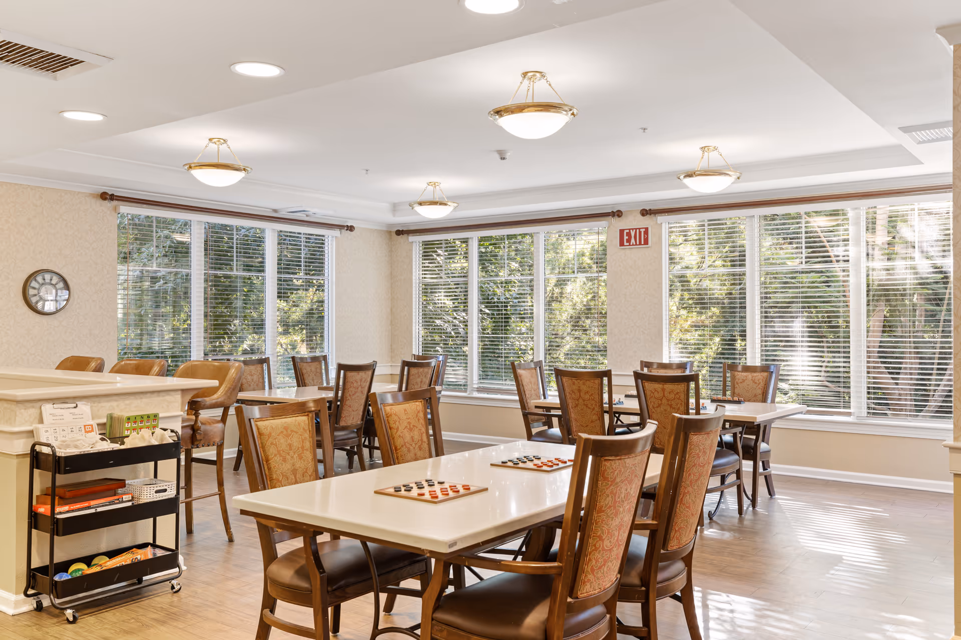 Bright communal dining/activity room with tables, chairs, large windows, and game boards on the tables.