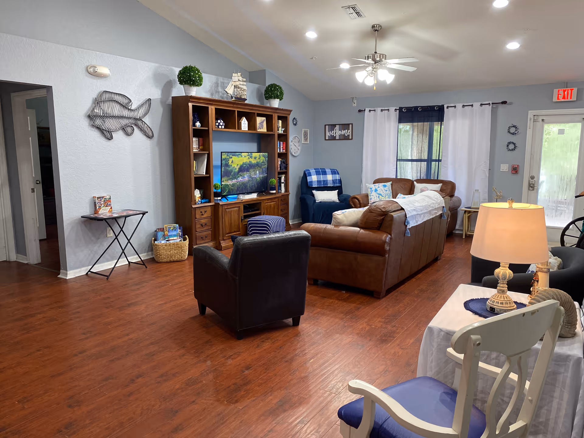 A cozy living room in Mariner Palms Assisted Living Facility featuring a brown leather sofa, a black armchair, a blue armchair with a plaid blanket, a wooden entertainment center with a TV, and various decorative items including plants and wall art. The room has wooden flooring, light blue walls, a ceiling fan with lights, and a window with white and blue curtains. A small table with a lamp and a white chair with a blue cushion are also visible.
