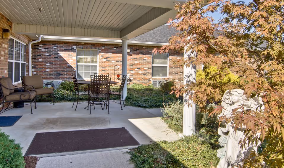 Covered outdoor patio area with a table and four chairs, two cushioned armchairs, surrounded by greenery and a brick building wall. A white angel statue is partially visible behind a tree with autumn-colored leaves.