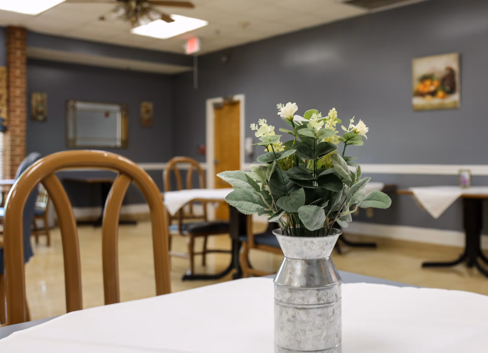 A dining area with wooden chairs and tables covered with white tablecloths. A metal vase with green and white artificial flowers is placed on the table in the foreground. The walls are painted gray with framed artwork and a mirror hanging on them.