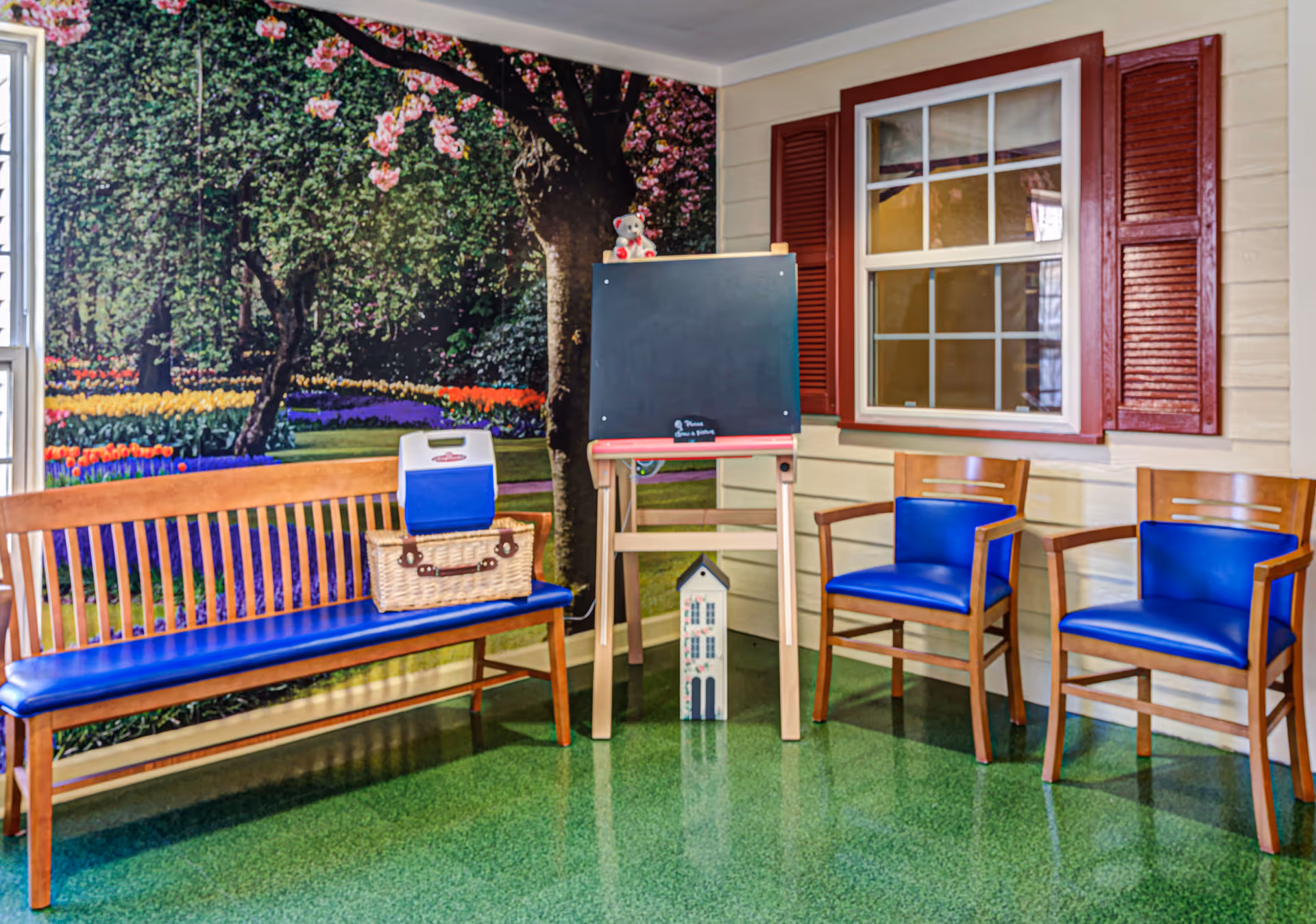 A cozy indoor seating area with a wooden bench and two wooden chairs, all with blue cushions. There is a small easel with a blackboard and a teddy bear on top. A picnic basket and a cooler are placed on the bench. The wall behind features a large mural of a garden with blooming flowers and trees, and a window with red shutters.