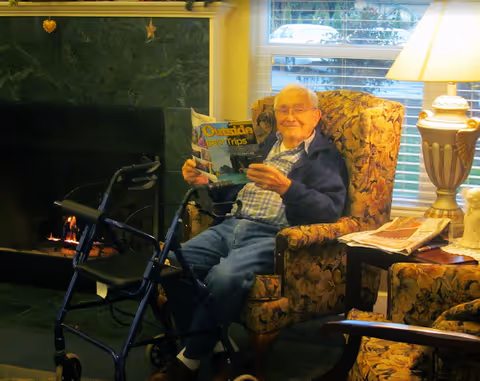 An elderly man sitting in a floral armchair next to a lit fireplace, holding a magazine titled 'Outside Best Trips'. A walker is positioned in front of him. There is a side table with a lamp and newspapers beside the chair, and a window with blinds in the background.