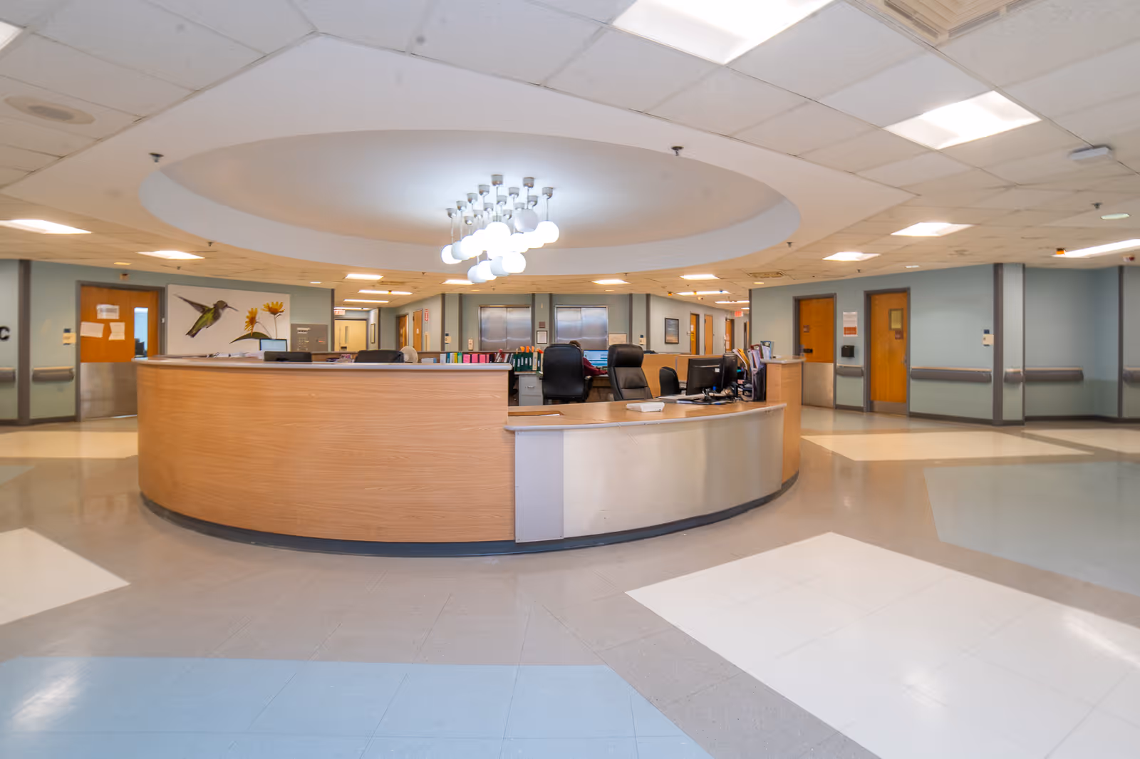 A spacious nursing station area inside a rehabilitation and nursing center with a large curved wooden desk, office chairs, computer monitors, and overhead lighting. The surrounding walls have doors and handrails, and there is a decorative painting of a hummingbird and flower on one wall.