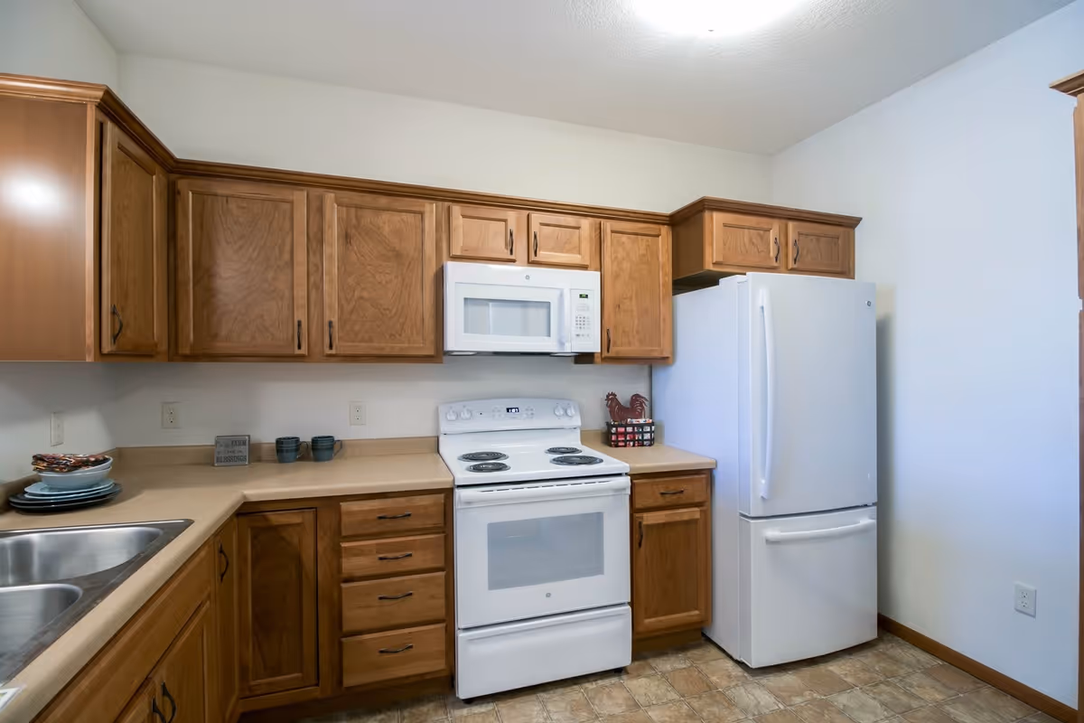 A kitchen with wooden cabinets, a white electric stove with oven, a white microwave above the stove, a white refrigerator, and a double stainless steel sink. The countertops are beige, and there are a few dishes and mugs on the counter.