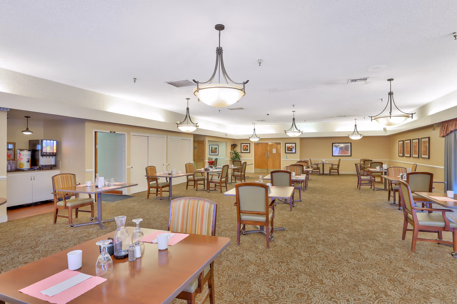 A spacious dining room with multiple tables and chairs arranged neatly. Each table has place settings including cups, glasses, and napkins. The room is well-lit with several hanging light fixtures and decorated with framed artwork on the walls. There is a beverage station with a drink dispenser on the left side near the entrance.