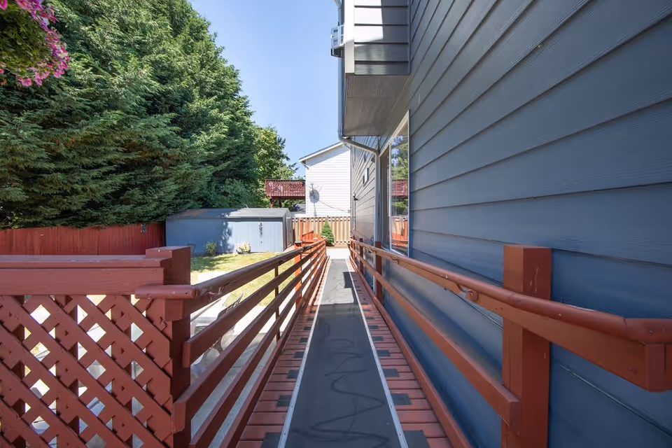 Outdoor view of a narrow walkway with red wooden railings on both sides leading to a fenced backyard area with a small shed. The right side of the walkway is bordered by a blue building wall, and tall green trees are visible on the left side under a clear blue sky.