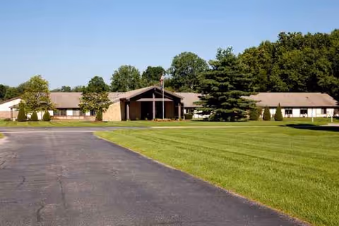 Single-story nursing facility building with a covered main entrance, flagpole, paved driveway, and a large manicured lawn.