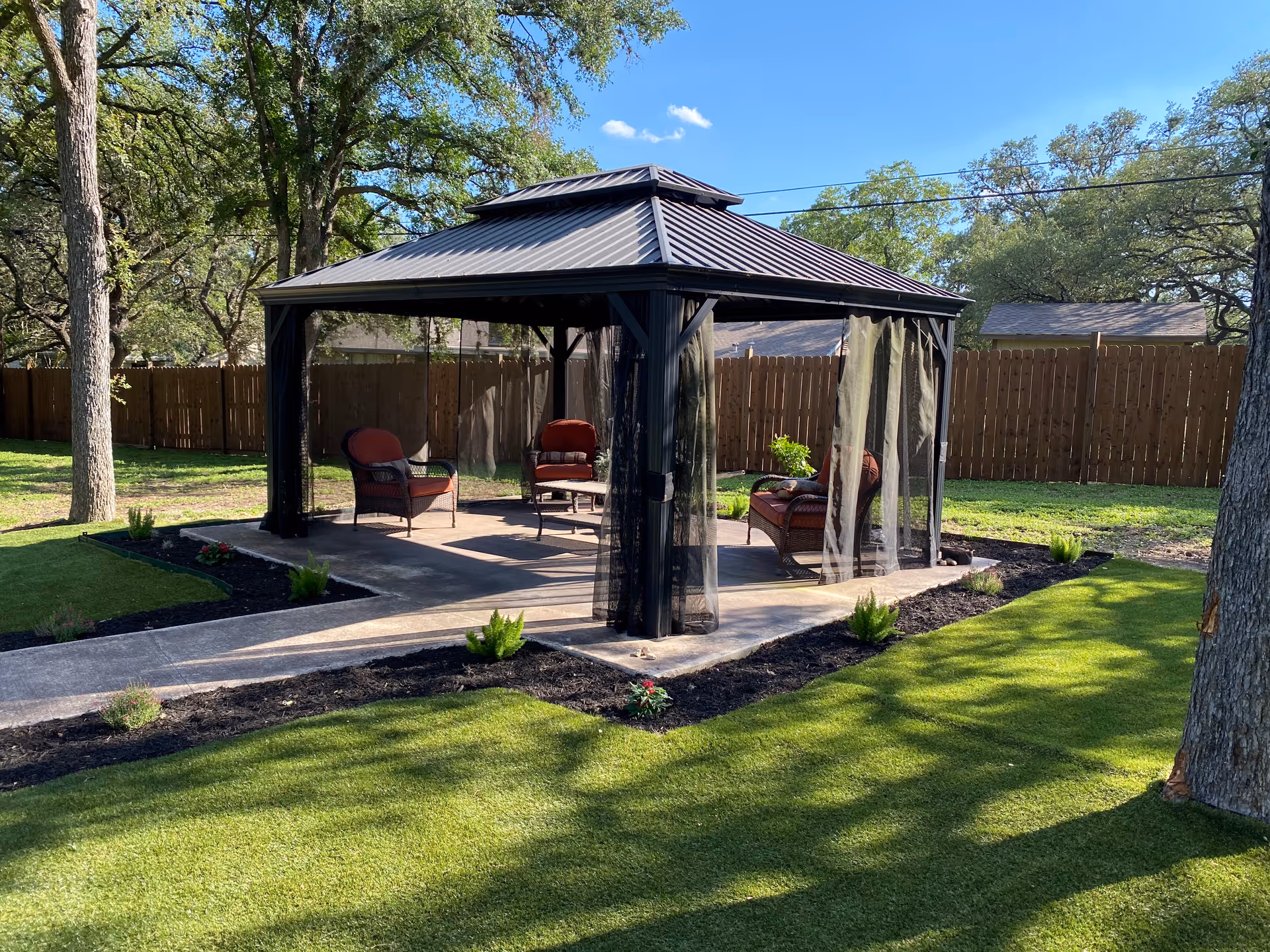 A covered gazebo with outdoor seating on a concrete patio in a grassy, tree-lined fenced yard.