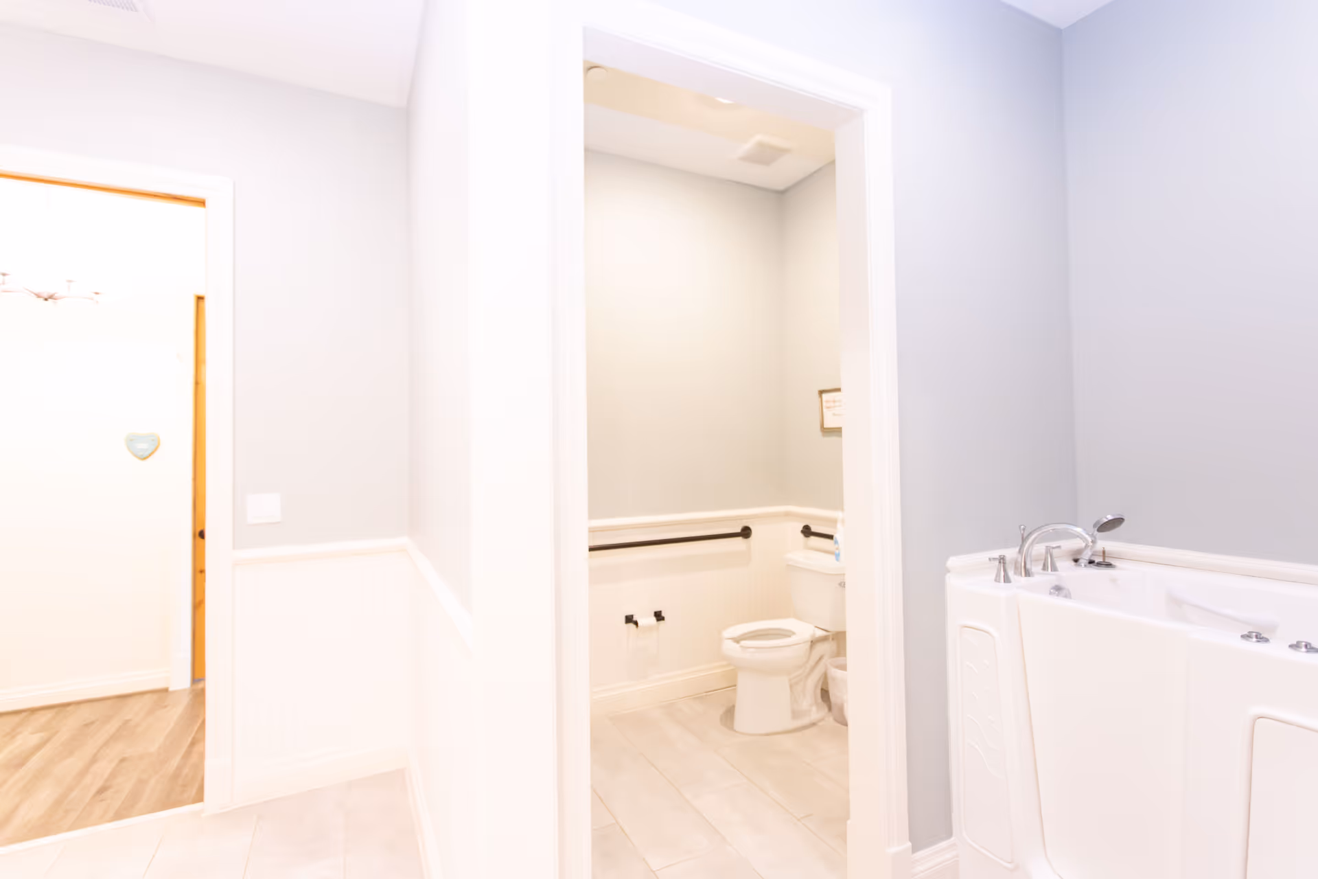 A clean and bright bathroom area in a senior living facility. The image shows a toilet with safety grab bars on the walls, a walk-in bathtub with faucet and handheld shower, light gray walls with white wainscoting, and tiled flooring. An open doorway leads to a hallway with wood flooring and a small heart-shaped wall decoration.