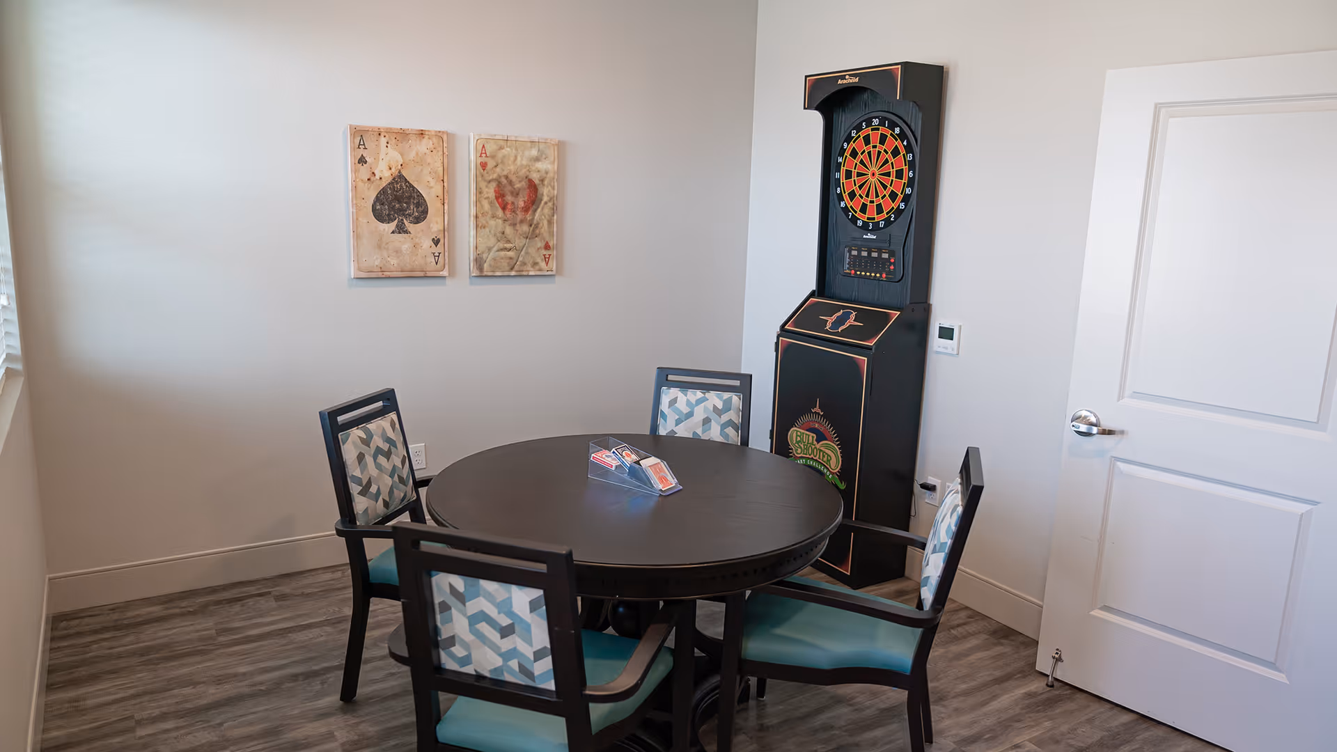 Small interior activity room with a round table surrounded by four chairs, a standing electronic dartboard, wall art, and a closed door.
