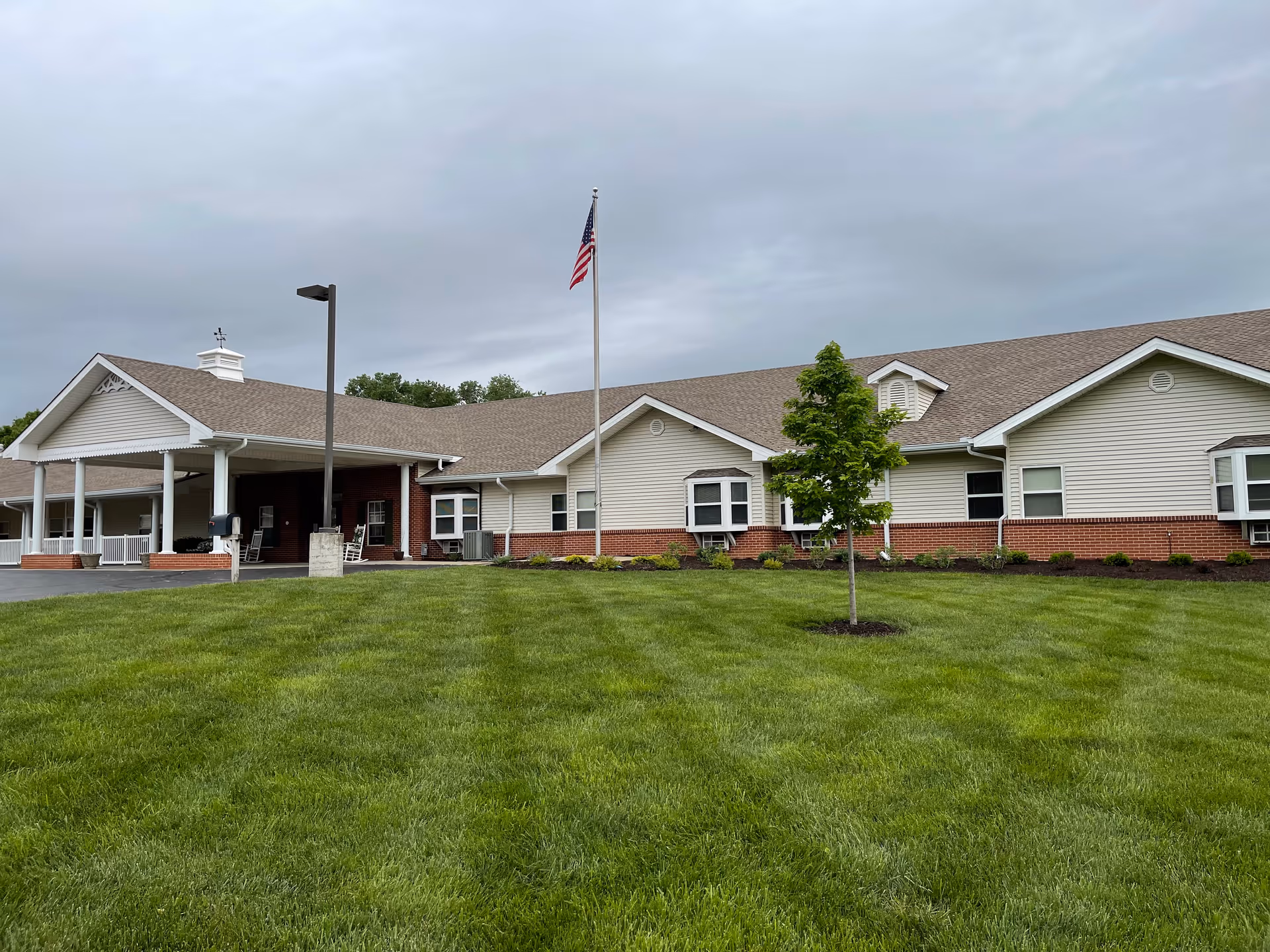 Single-story senior living building with a covered entrance, American flag, and a neatly mowed lawn under a cloudy sky.