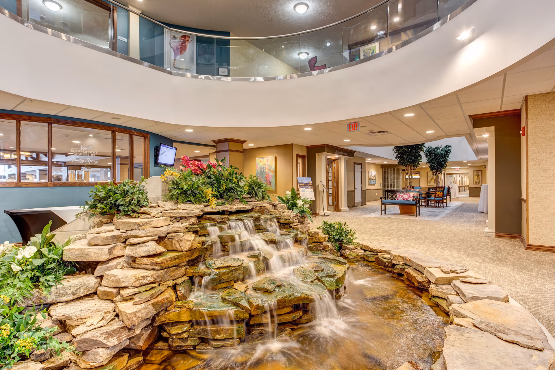 Atrium-style interior common area with a multi-tiered rock waterfall feature, plants, seating areas, and a curved upper balcony.