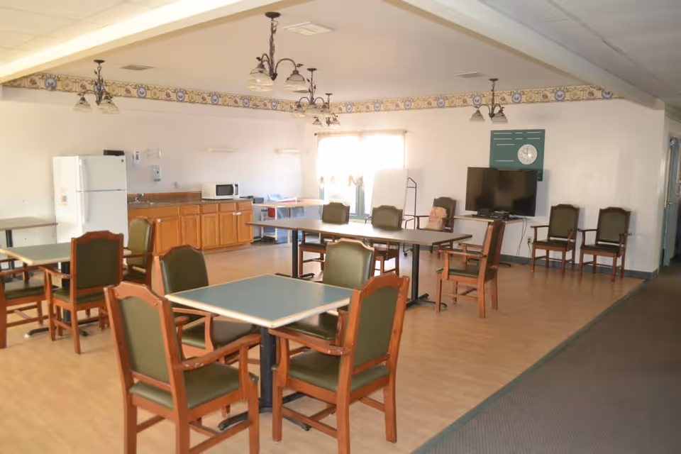 A spacious room with several tables and chairs arranged for dining or activities. The room features a kitchenette area with a refrigerator, microwave, and cabinets along one wall. There is a television mounted on the wall with additional chairs nearby. The room has wood flooring and is well-lit by natural light from a window with curtains.