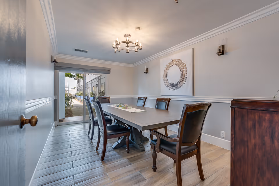 A dining room with a rectangular wooden table surrounded by six dark wooden chairs with black cushions. A modern chandelier hangs from the ceiling, and a large abstract circular artwork is mounted on the wall. The room has light-colored walls with white trim and wood-look tile flooring. A sliding glass door at the far end opens to an outdoor patio area with plants.