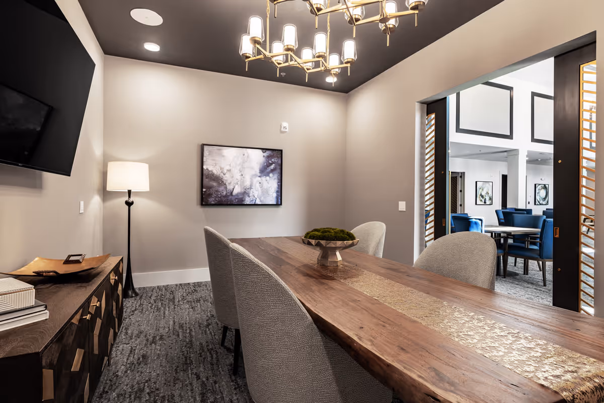 Contemporary dining/meeting room with a long wooden table, upholstered chairs, chandelier, and an adjoining seating area visible through an open doorway.