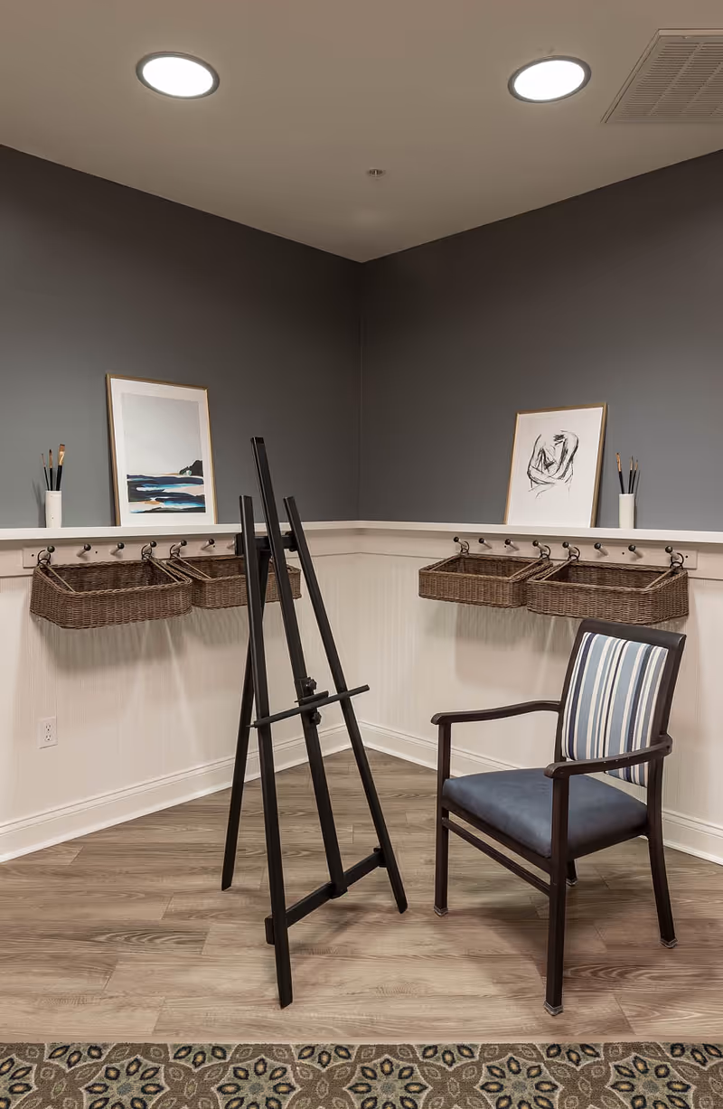 A corner of a room with gray walls and wood flooring, featuring a black easel, a wooden chair with blue and white striped upholstery, four wicker baskets hanging on hooks along the walls, two framed artworks resting on a shelf, and two white containers holding paintbrushes.