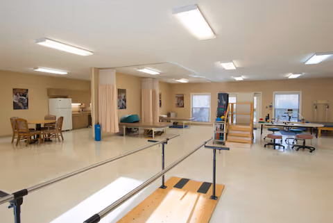 A spacious physical therapy or rehabilitation room with parallel bars for walking exercises, therapy tables, a small dining table with chairs, and various therapy equipment. The room has beige walls, large windows, and bright overhead lighting.