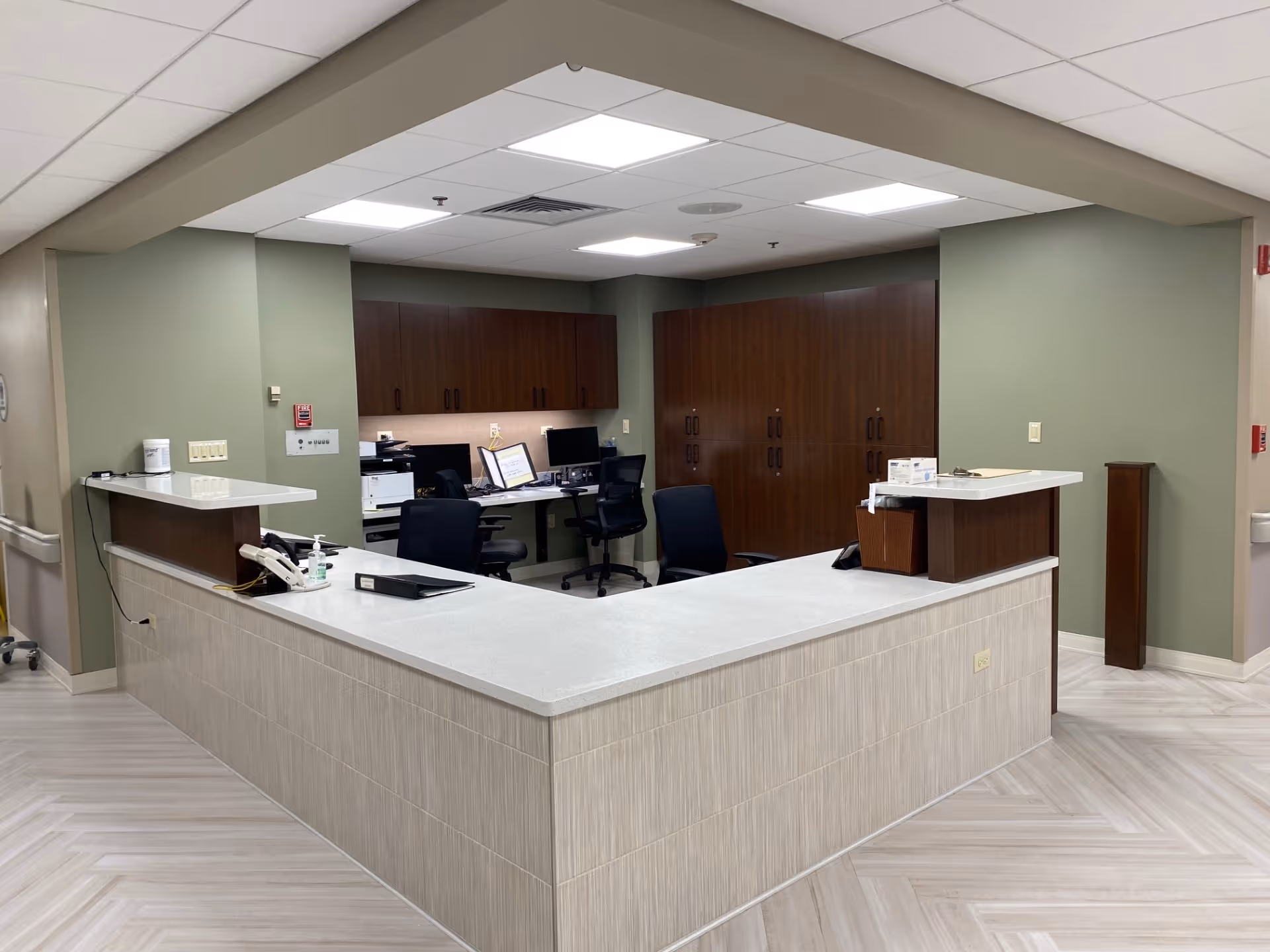 Reception or nurse station area inside St. Patrick's Residence Nursing and Rehabilitation with a large L-shaped counter, office chairs, computer monitors, and wooden cabinets against green walls.
